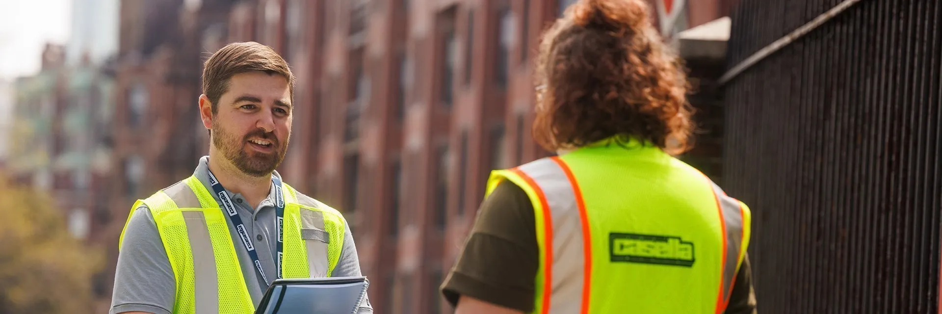 Two workers in high‑visibility vests standing beside Casella waste containers, reviewing information on a clipboard.