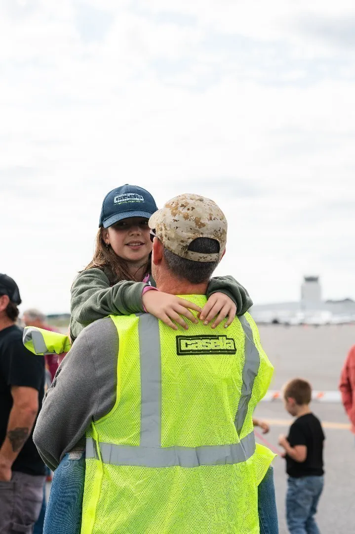 Casella employee and veteran wearing a high-visibility safety vest and camouflage cap holds a child.