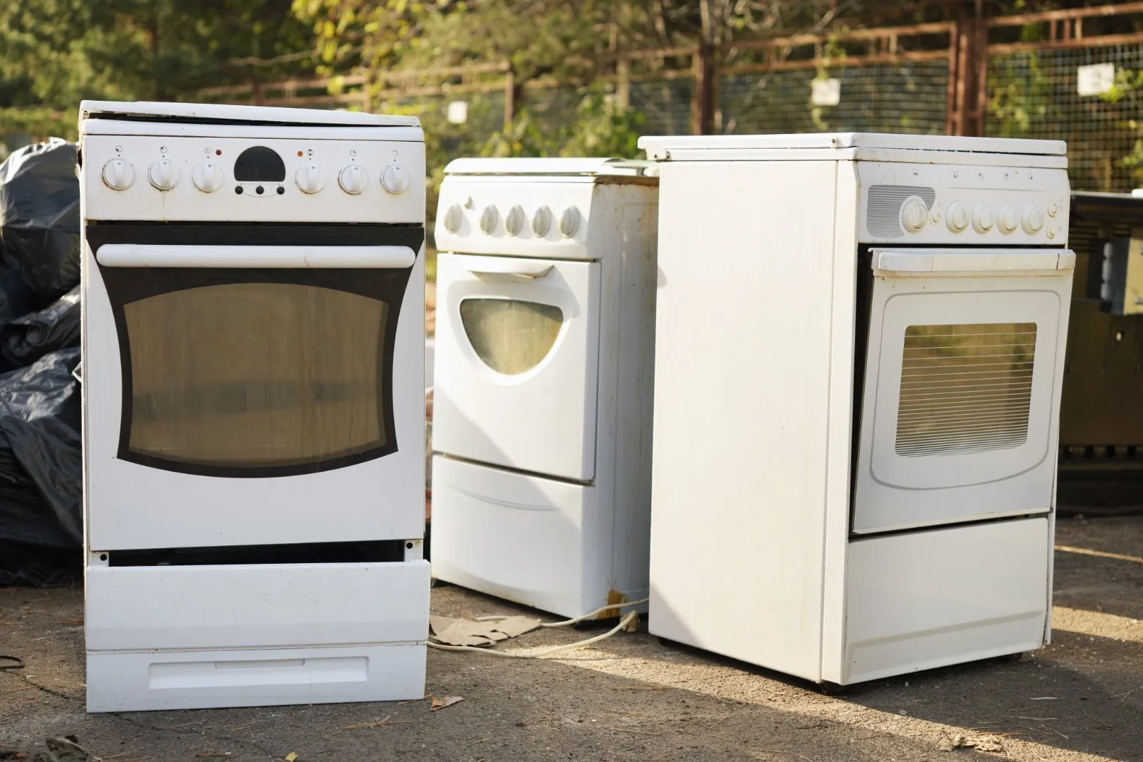 Three old white electric stoves with ovens placed outdoors on a paved surface, positioned for disposal or recycling. The appliances show signs of wear, including scratches and discoloration, and are arranged side by side near a wire fence with green foliage in the background.
