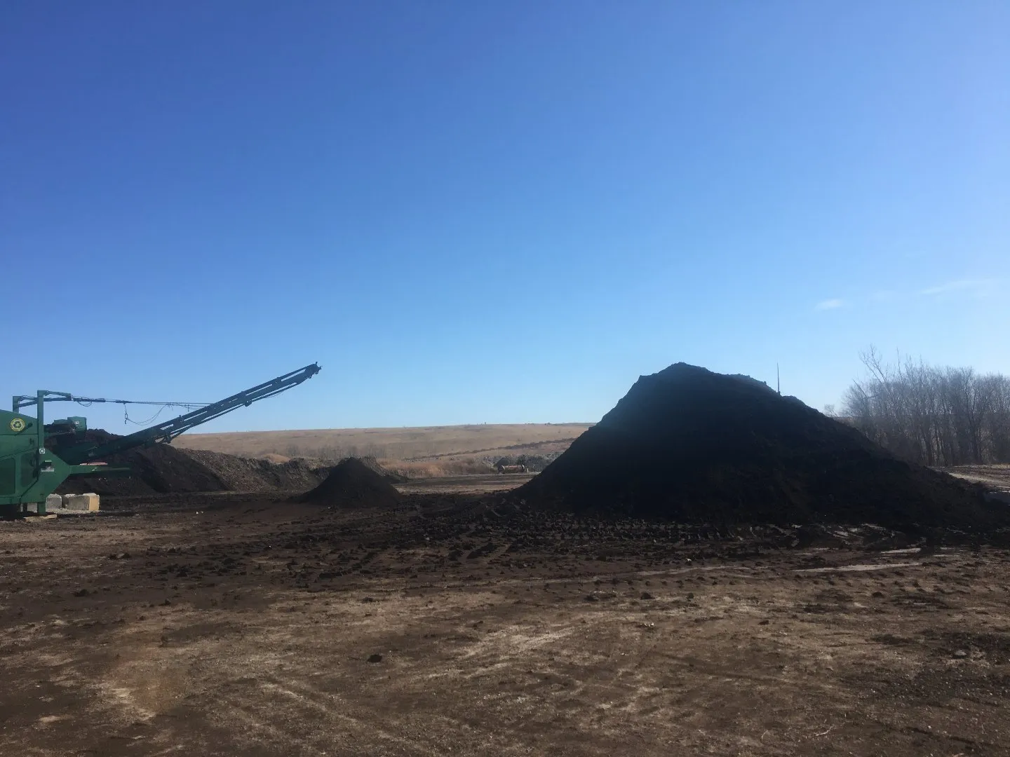 Compost processing site with a conveyor system and several large piles of dark compost material spread across an open, muddy area under a clear blue sky.