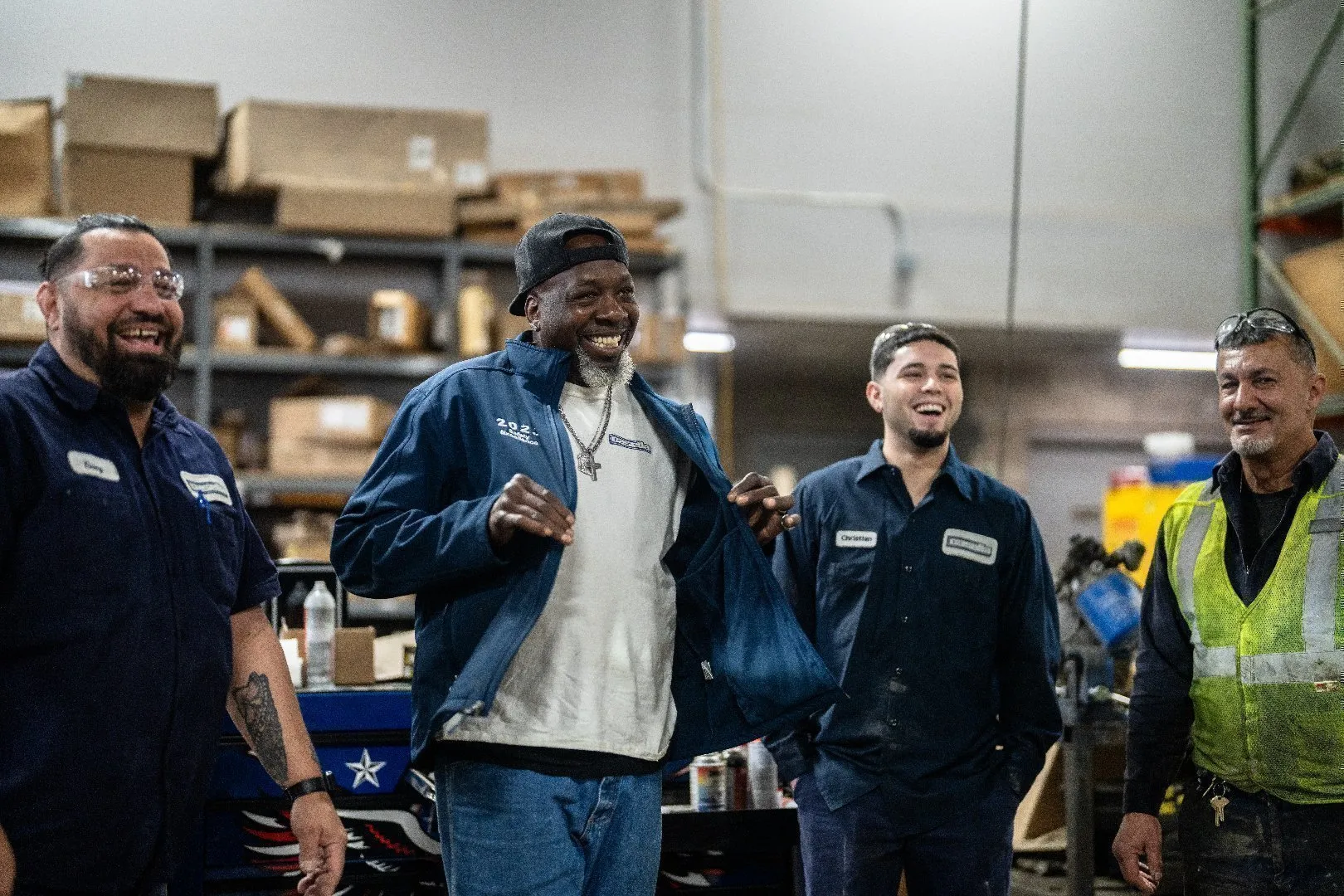 Four employees wearing work uniforms and safety gear stand together inside an industrial workshop with shelves of boxes and tools in the background.
