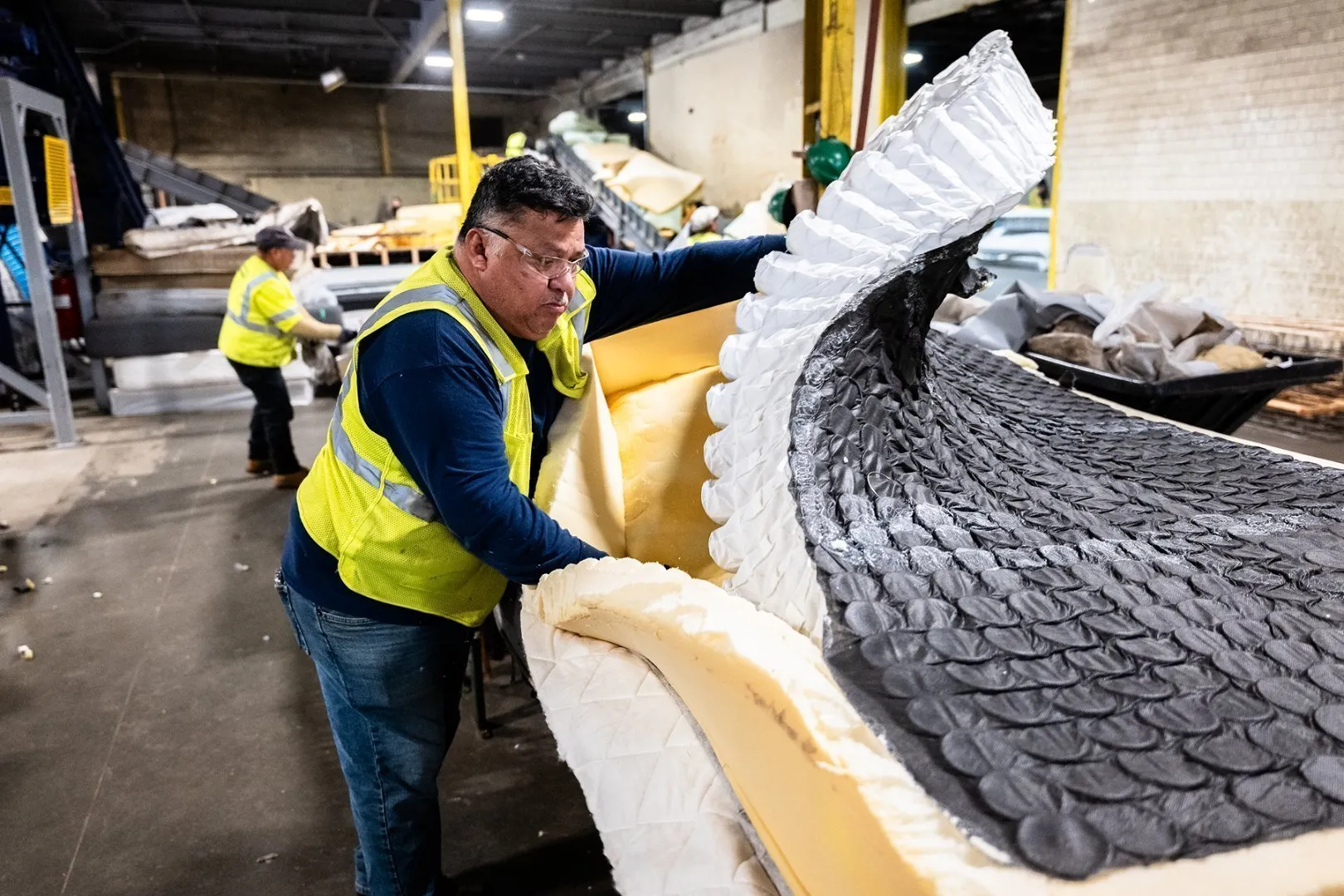 Worker wearing a bright yellow high-visibility safety vest dismantles a large mattress inside an industrial recycling facility, pulling apart foam and fabric layers. Additional mattresses and yellow support columns are visible in the background.