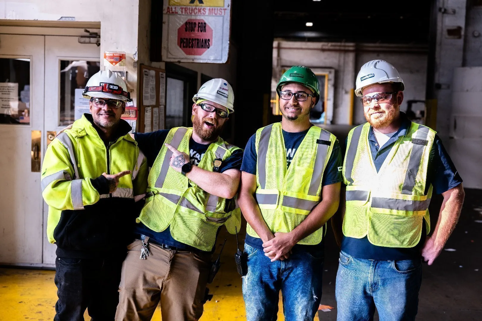 Four workers wearing high-visibility safety vests and hard hats stand together inside an industrial facility.