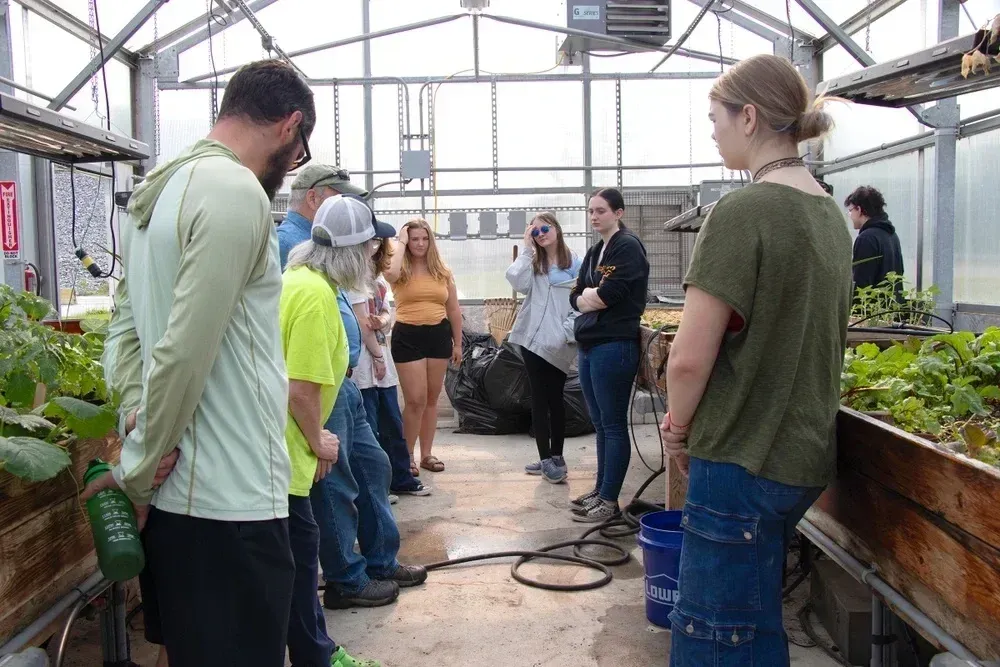 Group of people standing inside a greenhouse during a discussion or educational session. Wooden planting beds filled with green leafy plants line both sides of the walkway, and gardening tools such as a hose and buckets are visible on the floor.