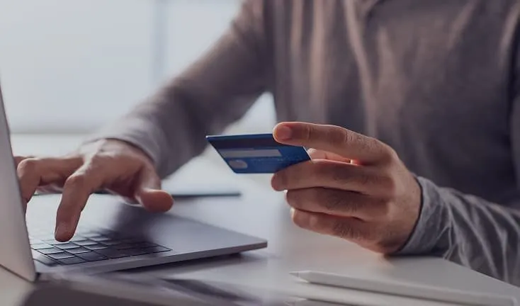Person holding a blue credit card while typing on a laptop keyboard, illustrating online payment or e-commerce transaction on a clean white desk.