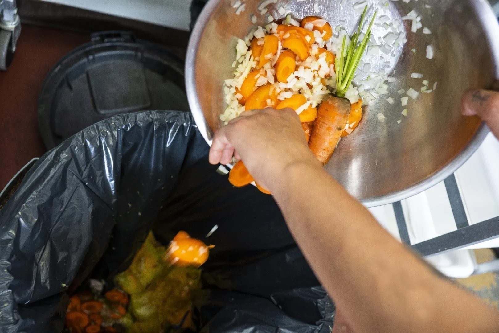 Person holds a large metal bowl filled with chopped carrots, onions, and vegetable stems, tipping the contents into a black plastic-lined compost bin with a round lid. Additional food scraps are visible inside the bin, and the setting appears to be a kitchen or food preparation area.