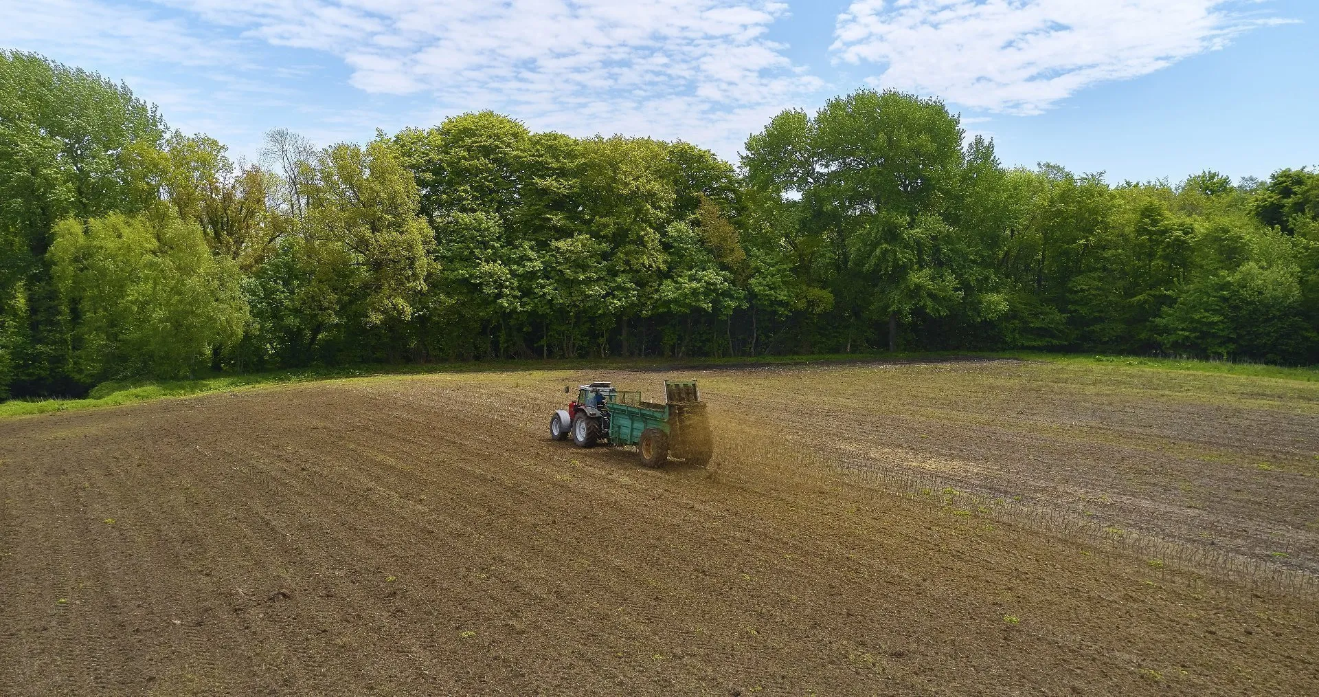 Tractor pulling a spreader that applies organic material across a large, recently tilled field, with a tree line and partly cloudy sky in the background.