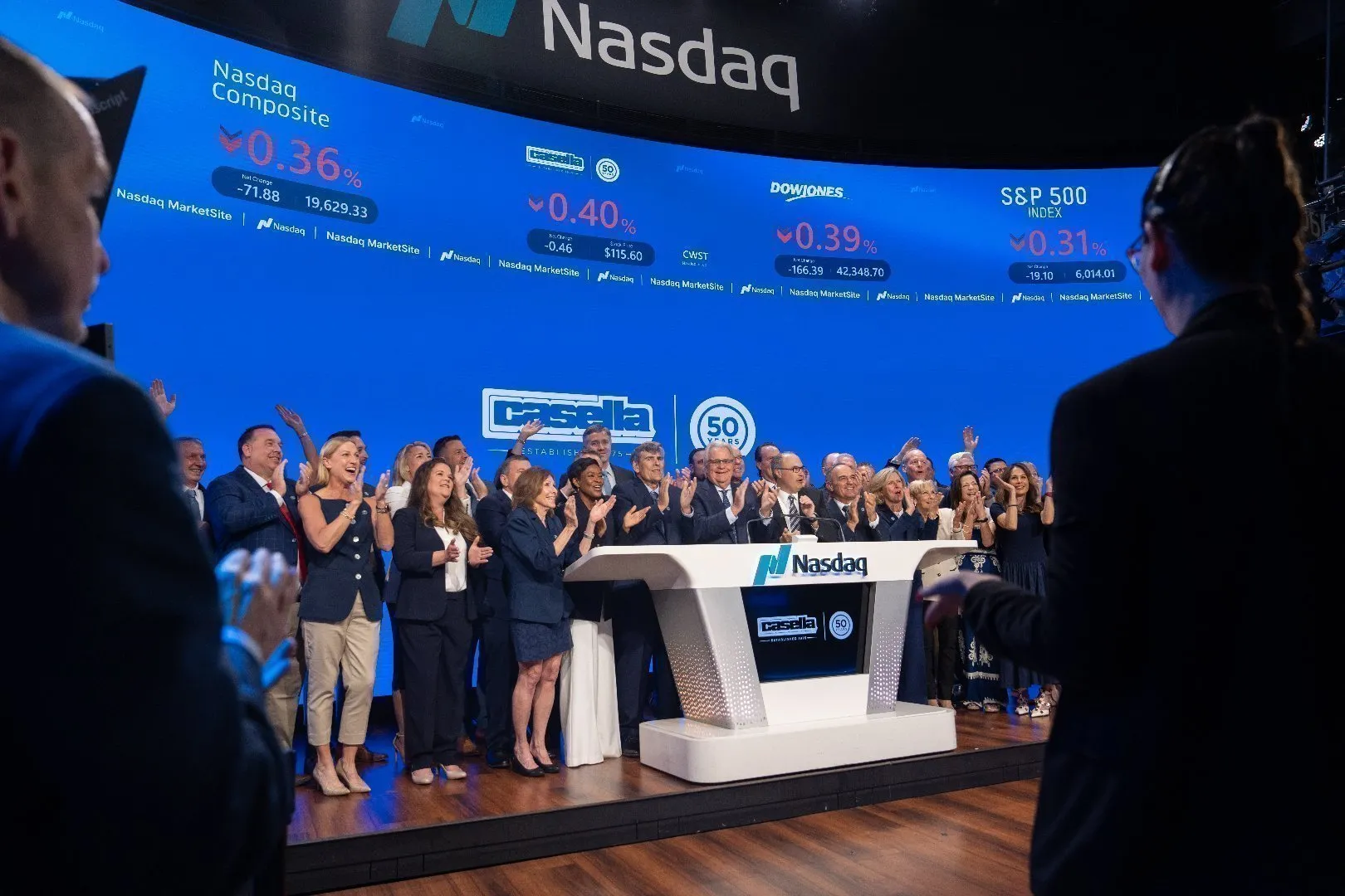 Casella team gathered at the Nasdaq opening bell podium, dressed in formal business attire, during a 50th anniversary celebration, clapping and smiling as the company logo and Nasdaq branding appear on a large blue digital display showing market indices.