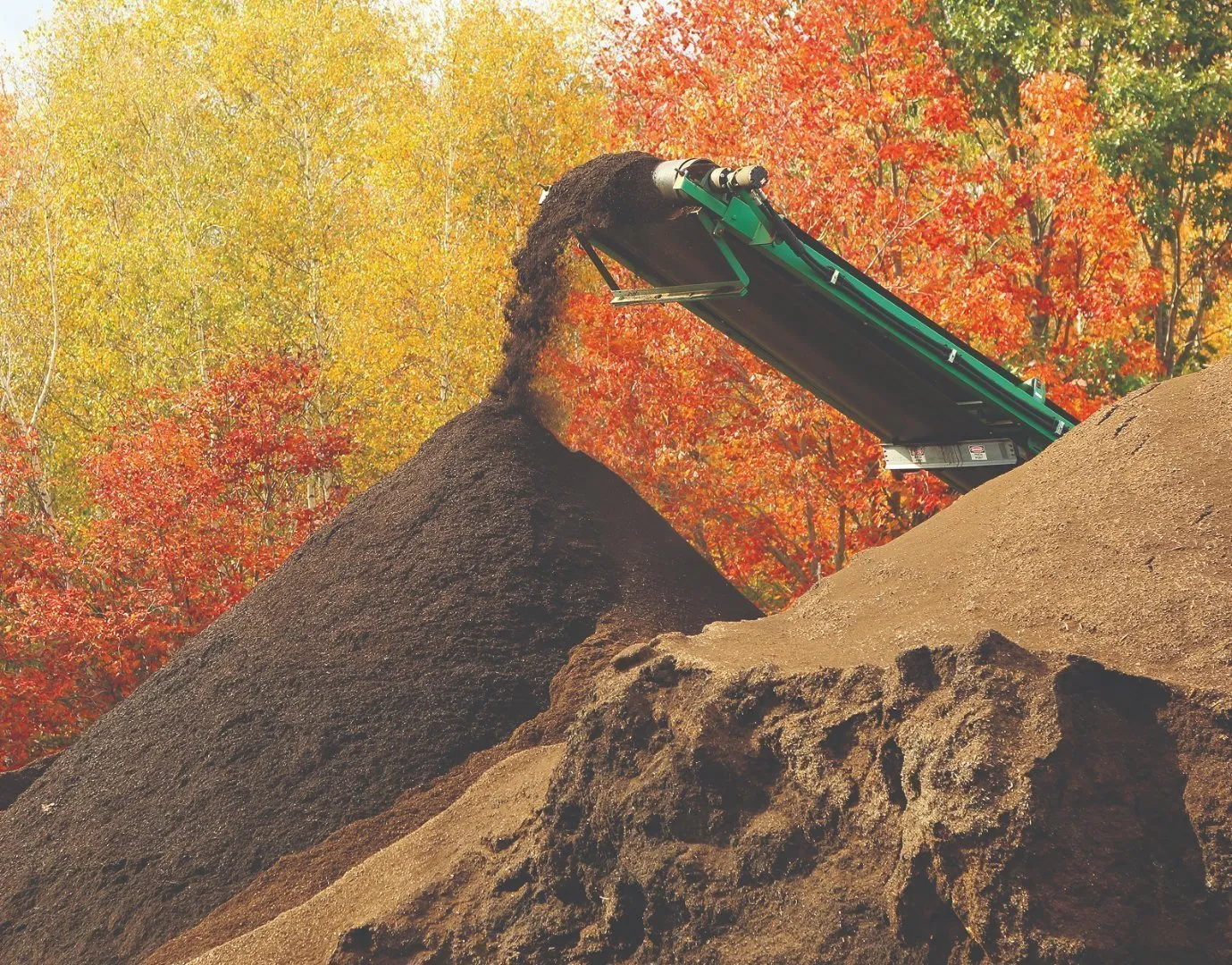 Green conveyor belt releases a stream of dark compost material onto a large pile at an outdoor organics processing site. Surrounding trees display vibrant autumn foliage in shades of red, orange, and yellow under bright daylight.