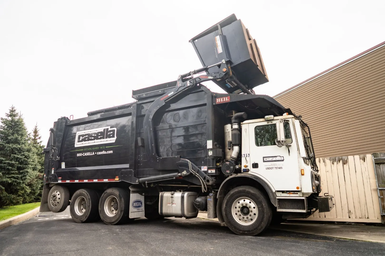 Casella front-loading garbage truck lifts a large commercial dumpster overhead in a paved service area next to a beige building with wooden fencing.