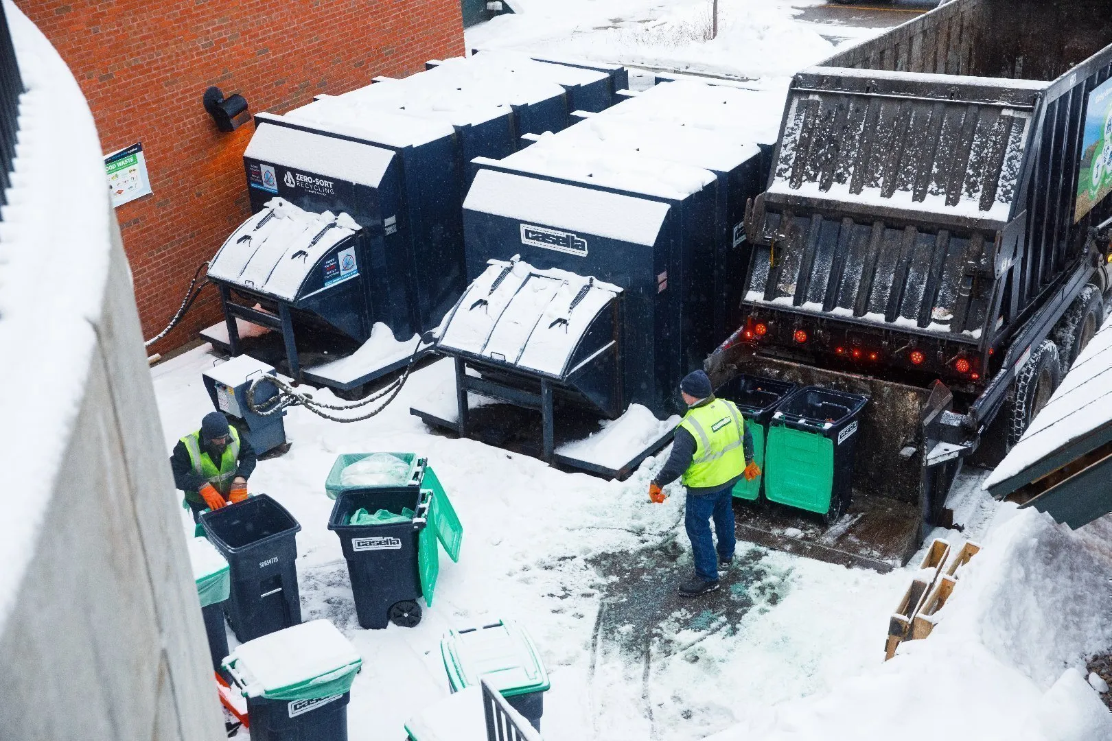 Green waste collection truck equipped with an automated arm lifts a small green curbside bin overhead for emptying. A worker wearing a bright yellow high-visibility safety vest and gloves stands nearby on a wet pavement next to a white utility vehicle.