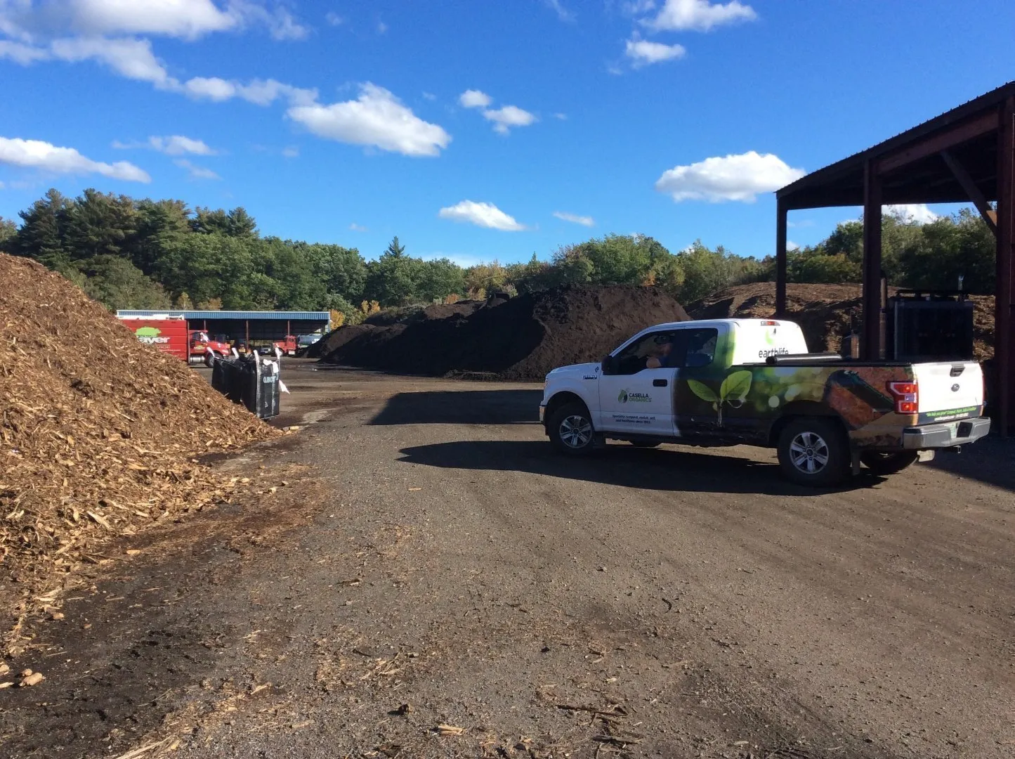 White pickup truck with Casella Organics branding parked at an outdoor composting facility. Large piles of mulch and compost material are visible on both sides of the dirt roadway, with a covered structure on the right and red equipment in the background.