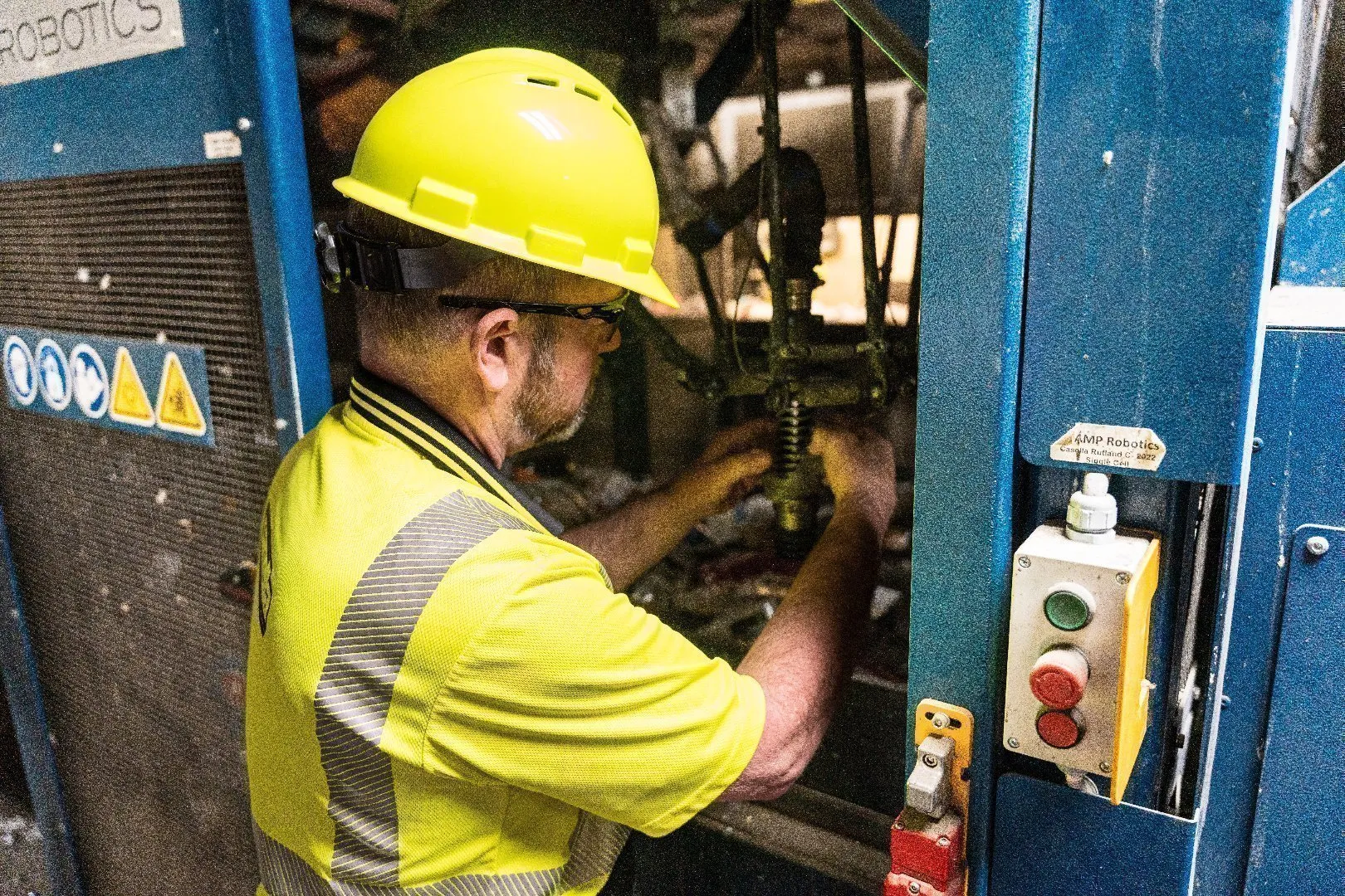 Technician wearing a yellow hard hat and high-visibility safety shirt performs maintenance inside a blue industrial machine.