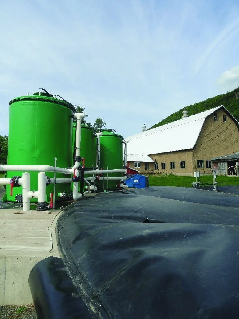 Green cylindrical tanks connected by white pipes and valves at an outdoor anaerobic digestion system near a farm building with a white roof. A large black geomembrane cover is visible in the foreground, and grassy hills with trees form the background under a partly cloudy sky.