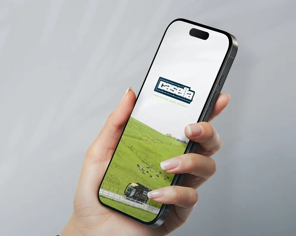 Hand holding a smartphone displaying the Casella logo on the screen above a green grassy landscape with a blue sky background, representing mobile access to Casella services. 