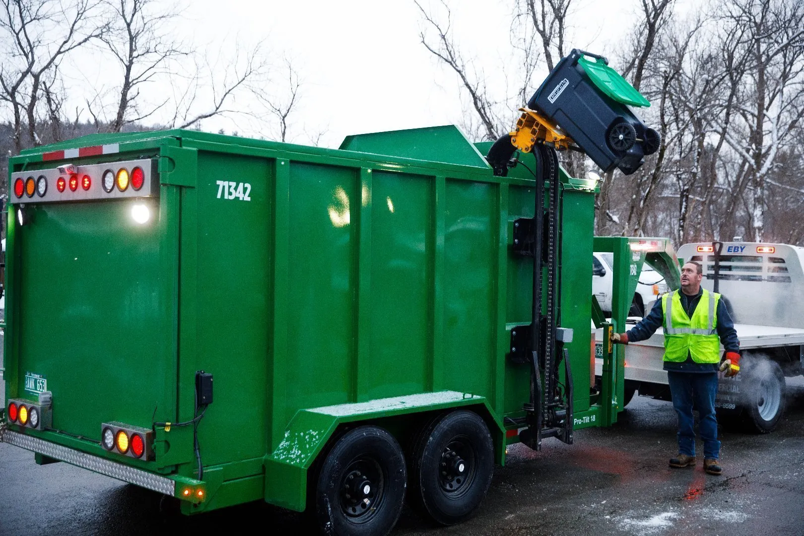 Two workers wearing bright yellow high-visibility safety vests and gloves manage green Casella organics carts in a snowy outdoor area next to a row of black food waste collection units. A large black truck with an open rear compactor is positioned for loading, and snow covers the ground and equipment.