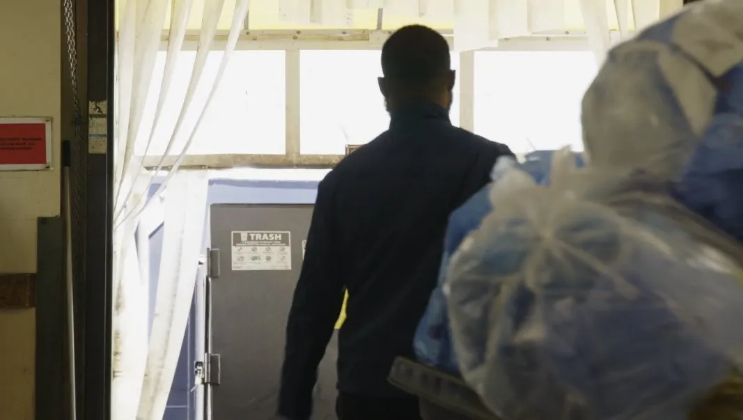 Person wearing a dark jacket walks toward a gray trash compactor labeled “TRASH” inside a facility. Large clear and blue plastic bags filled with medical materials are stacked on a cart in the foreground.