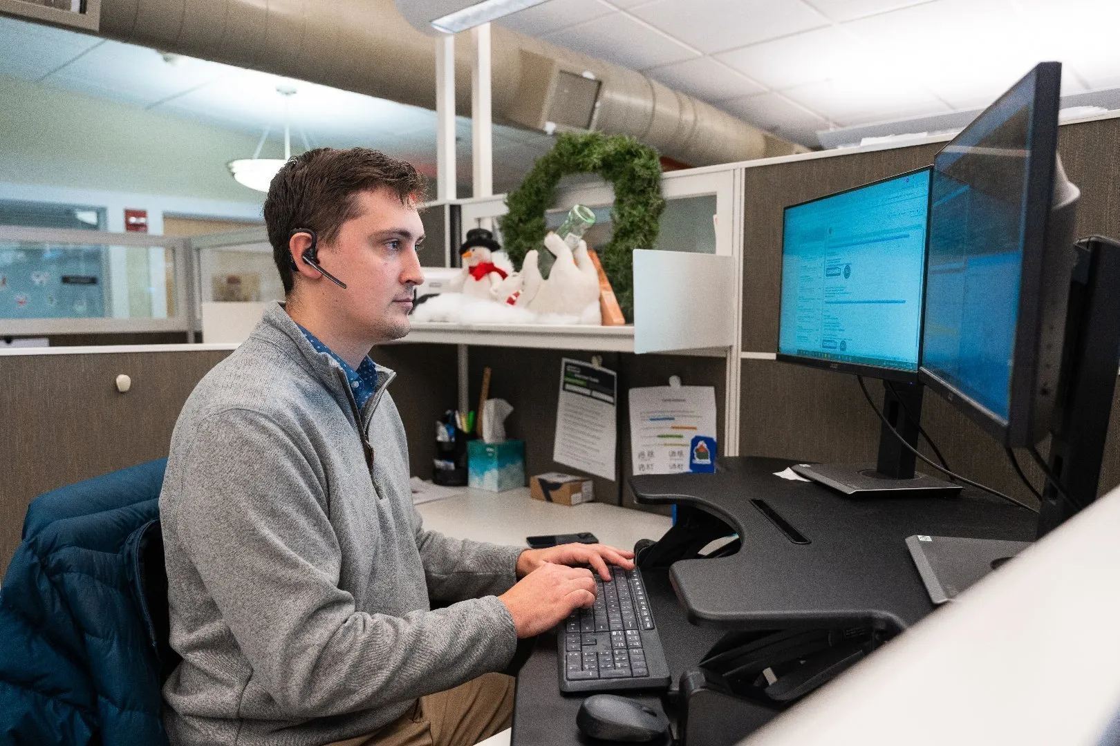 Accounts Receivable Specialst seated at an office cubicle wearing a gray sweater and using a dual-monitor computer setup with a keyboard and mouse.