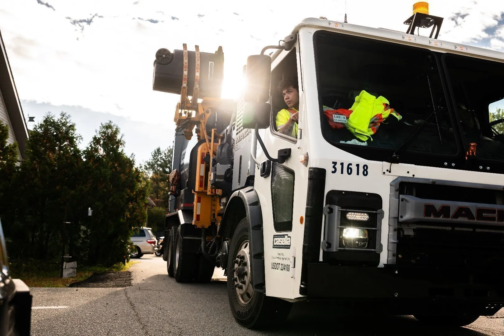 Casella automated side-load garbage truck on a residential street with sunlight shining through, featuring a mechanical arm and high-visibility safety gear inside the cab.