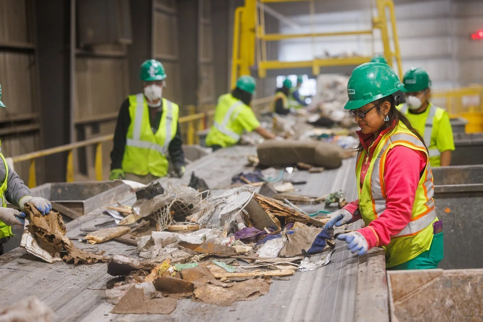 Workers wearing green hard hats, bright yellow high-visibility safety vests, and gloves sort mixed recyclable materials on a conveyor belt inside a material recovery facility. The sorting line includes paper, cardboard, and other debris, with yellow safety railings and industrial equipment visible in the background under bright overhead lighting. 