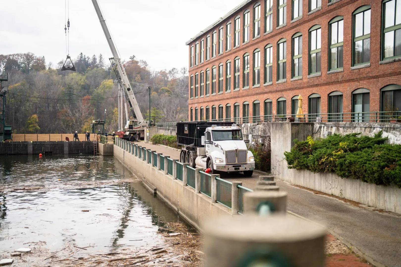 White semi-truck with a black roll-off dumpster parked on a narrow roadway beside a large red brick industrial building. A crane with a suspended load operates near a waterway bordered by concrete walls and green railings.