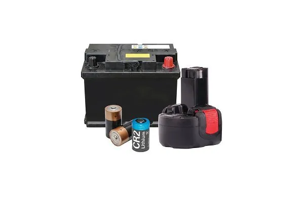 Collection of batteries with a red prohibition symbol over them, indicating they are not allowed for recycling. Items include a large black car battery with red and black terminals, two cylindrical batteries, a blue CR2 lithium battery, and a black cordless power tool battery pack. 