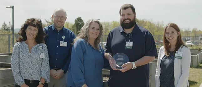 Five adults standing outdoors, smiling, as a bearded man holds a glass award.