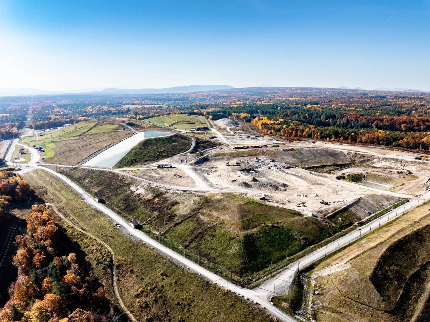 Aerial view of a landfill with waste cells, access roads, a leachate pond, heavy equipment, and autumn-colored trees under a clear blue sky.