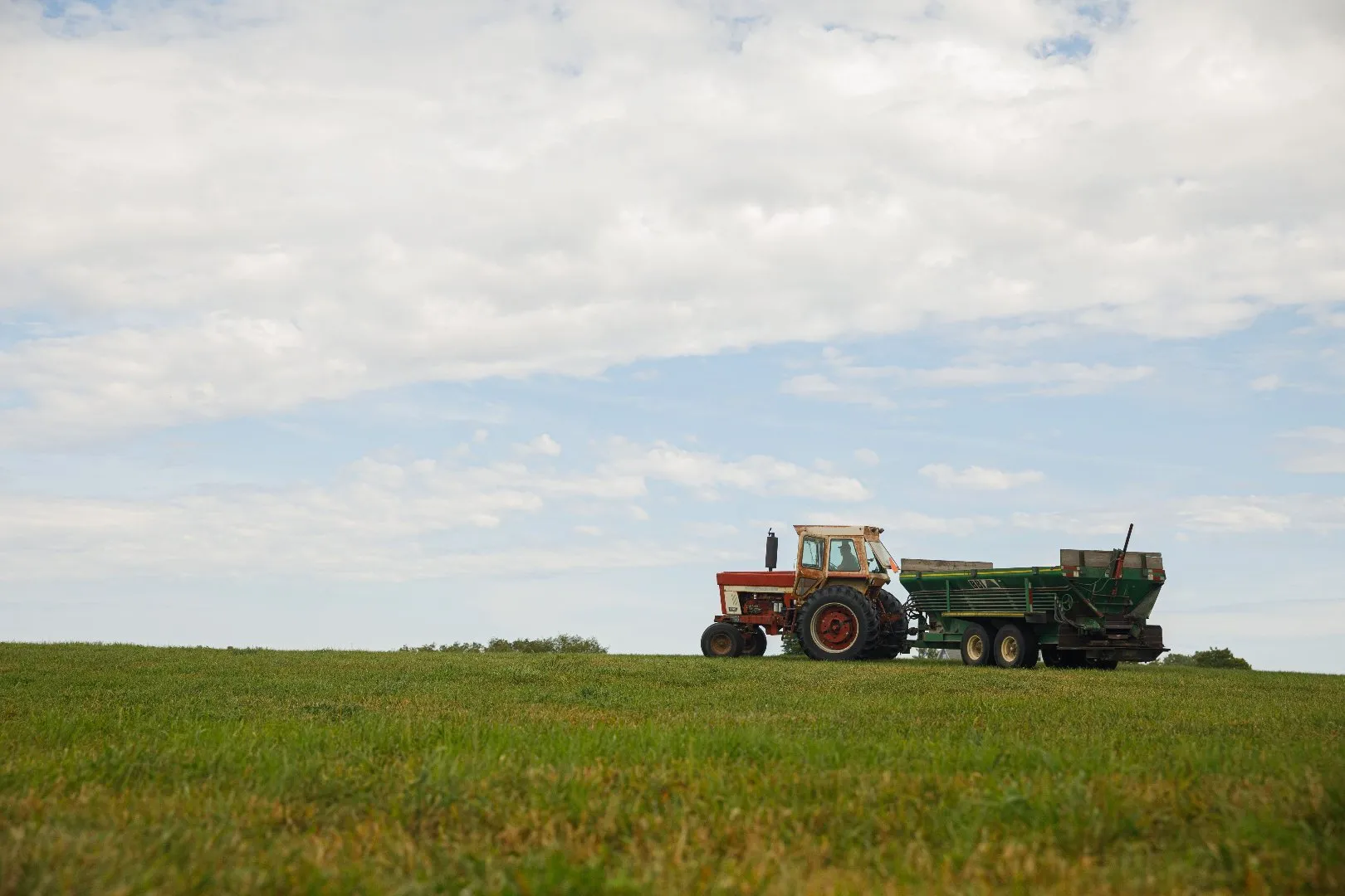 Tractor pulling a green spreader across a grassy field under a wide sky with large white clouds.