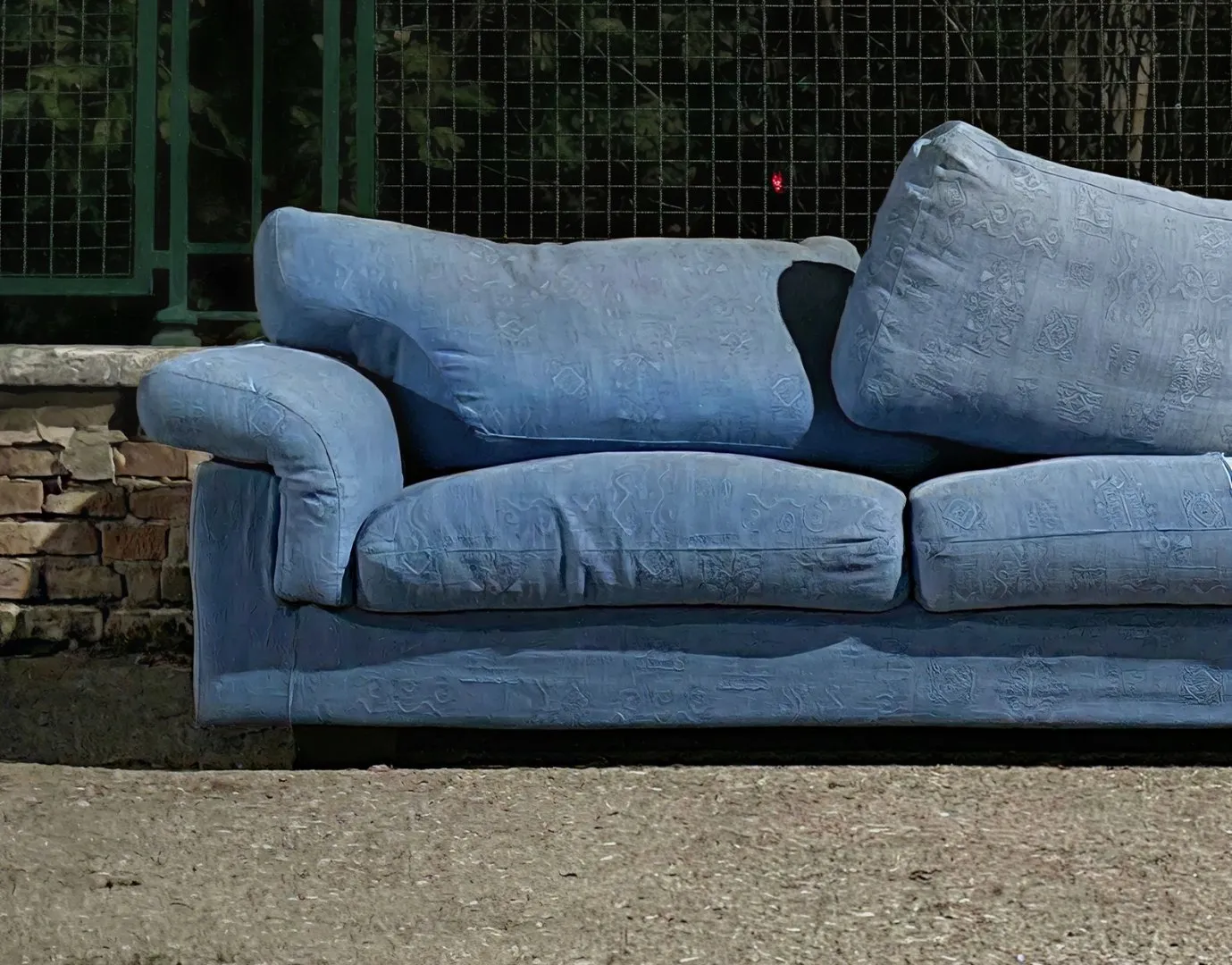 Blue upholstered sofa with three seat cushions and three backrest cushions sits outdoors, discarded on a sidewalk near a metal fence and brick wall.