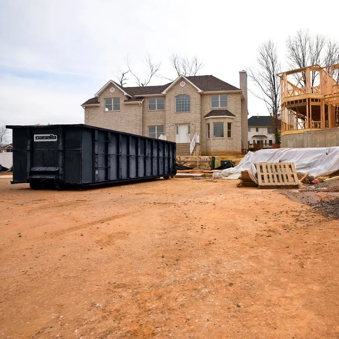 Large Casella roll-off dumpster placed on a dirt construction site in front of a newly built beige two-story house with dark brown roof shingles. To the right, a wooden-framed structure is under construction, and several wooden pallets and plastic-covered building materials are scattered across the ground.