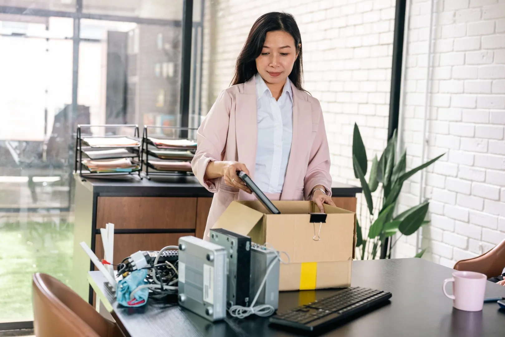 Person wearing a light pink blazer and white shirt packs office items into a cardboard box on a desk. The desk contains a keyboard, phone, cables, and other electronic accessories, with a stack of file trays and a potted plant visible in the background. The setting features white brick walls and large glass windows.