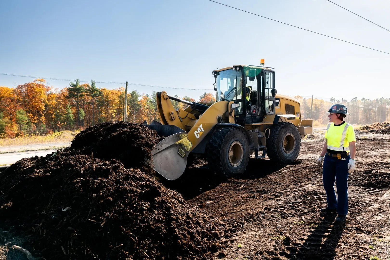 Yellow CAT front loader moves a large pile of dark compost material at an outdoor organics processing site. A worker wearing a bright yellow high-visibility safety shirt, hard hat, and gloves stands nearby observing the operation. Autumn-colored trees and a clear blue sky form the background.