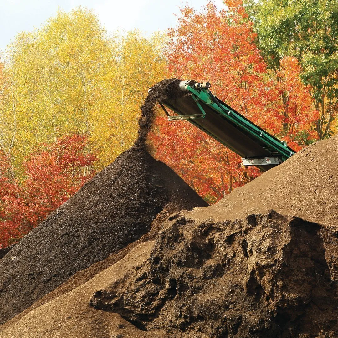 Green conveyor belt deposits compost onto a large pile at an organics processing site, with colorful autumn trees in the background.