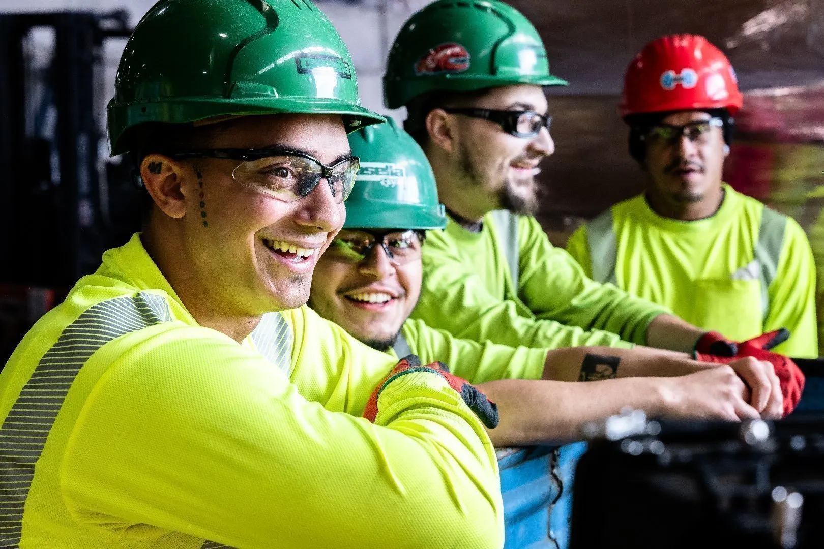 Workers wearing green and red hard hats and bright yellow high-visibility safety shirts stand side by side near industrial equipment inside a facility.
