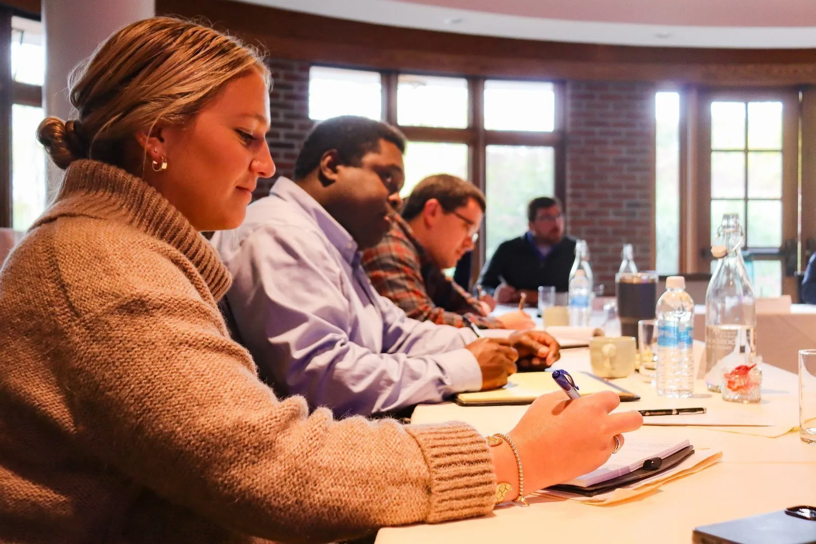 Group of people seated at a conference table in a meeting room with large windows and brick walls, taking notes on notepads.
