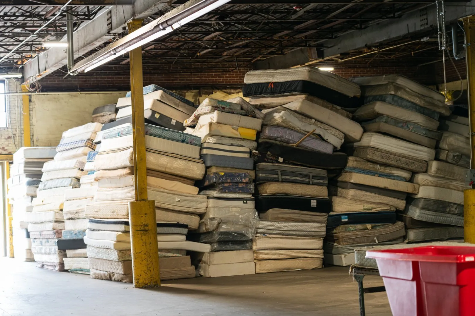 Large stacks of used mattresses piled high inside an industrial warehouse with exposed beams and yellow support columns, ready for recycling. A red plastic bin is visible in the foreground.
