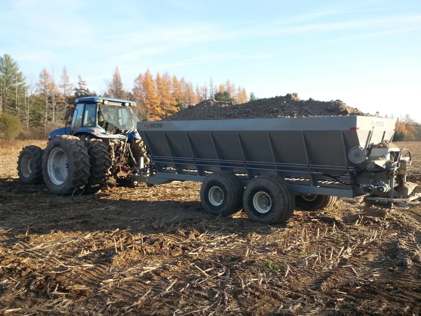 Blue tractor pulling a spreader filled with soil or compost across a harvested field, with trees and a clear sky in the background.