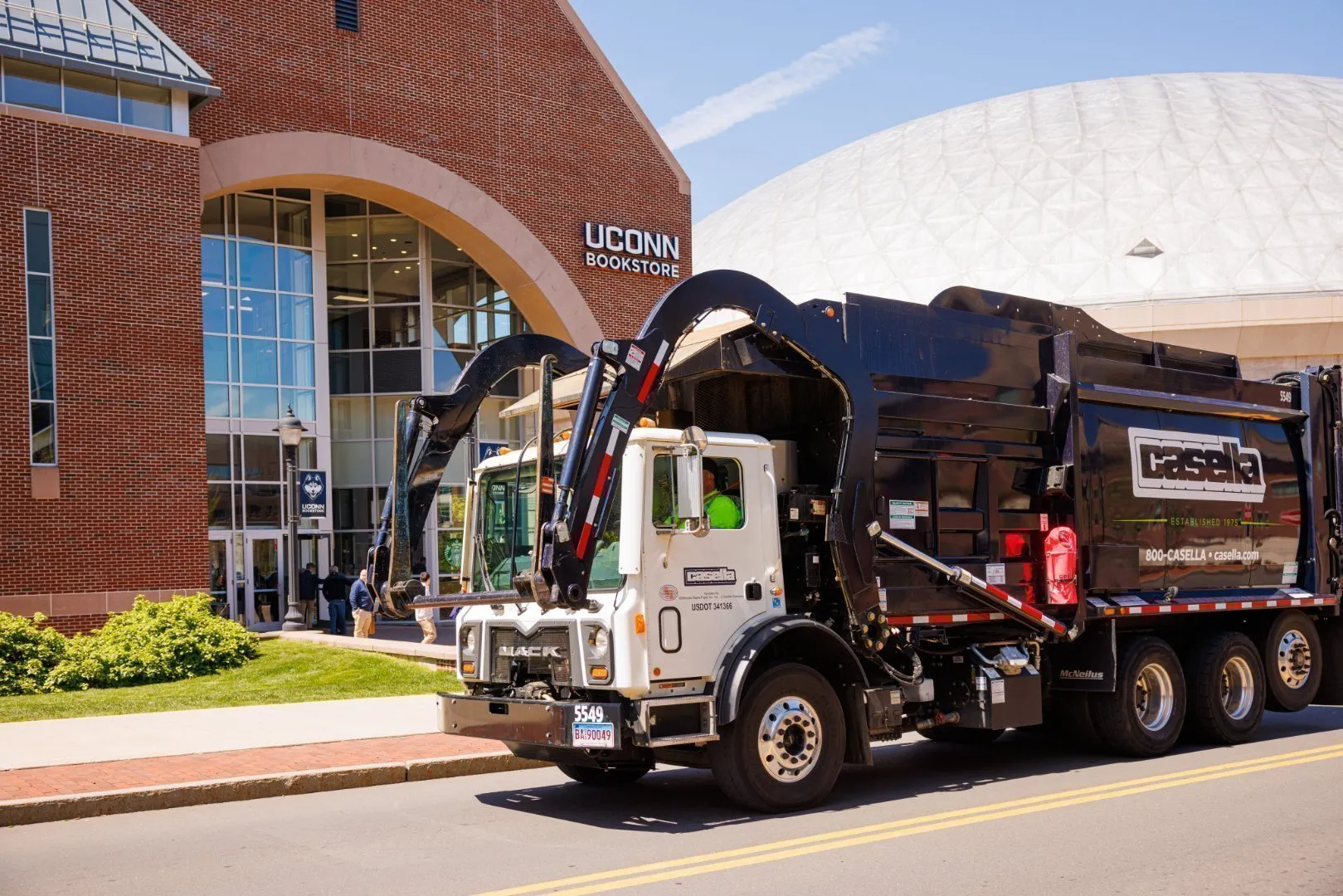 Casella waste collection truck parked on a street in front of the UConn Bookstore and a large white domed building on the University of Connecticut campus. The truck features a front-loading arm and Casella branding, while the background includes brick architecture, glass entrance doors, and campus signage under a clear blue sky.