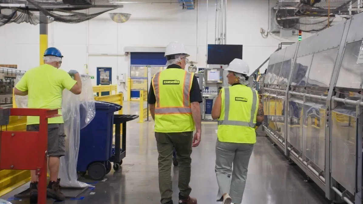 Two workers wearing white hard hats and bright yellow high-visibility safety vests walk through an industrial facility next to a large automated sorting machine. Another worker in a blue hard hat and neon yellow shirt stands nearby handling a clear plastic bag. The setting includes polished concrete floors, yellow safety railings, blue bins, and overhead equipment under bright lighting.