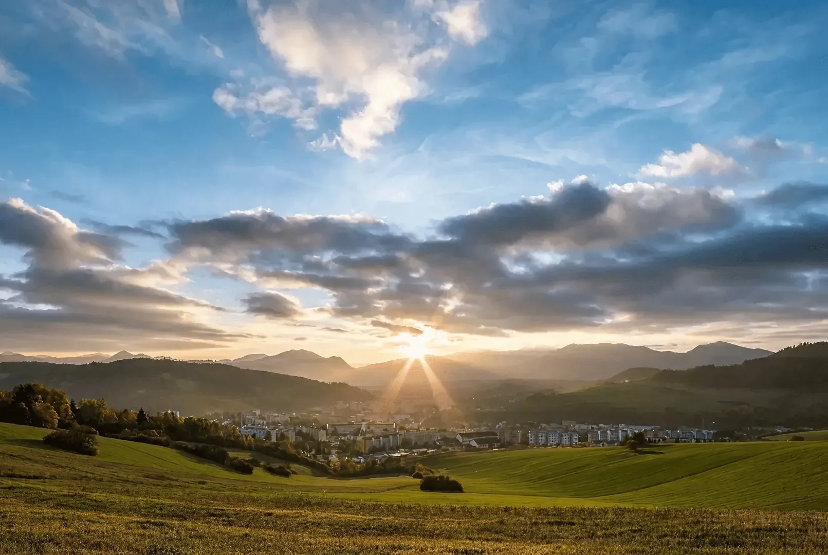 Sun rays over a small town in a green valley with hills and mountains.