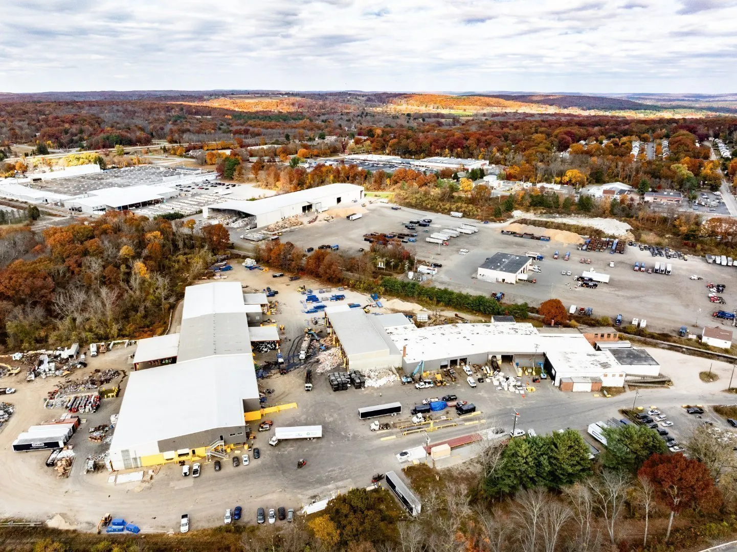 Aerial view of a large Casella recycling facility surrounded by parking areas, trucks, and autumn-colored trees, with distant hills under a partly cloudy sky.