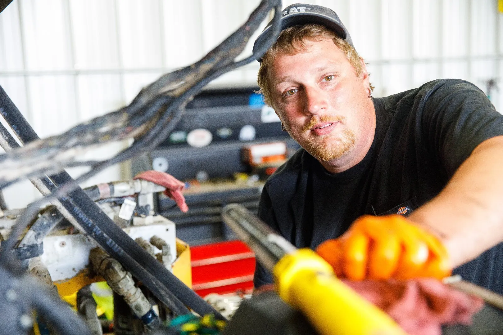 Equipment technician wearing orange protective gloves works on hydraulic components of heavy equipment inside a workshop, with visible hoses, metal parts, and a red tool chest in the background.