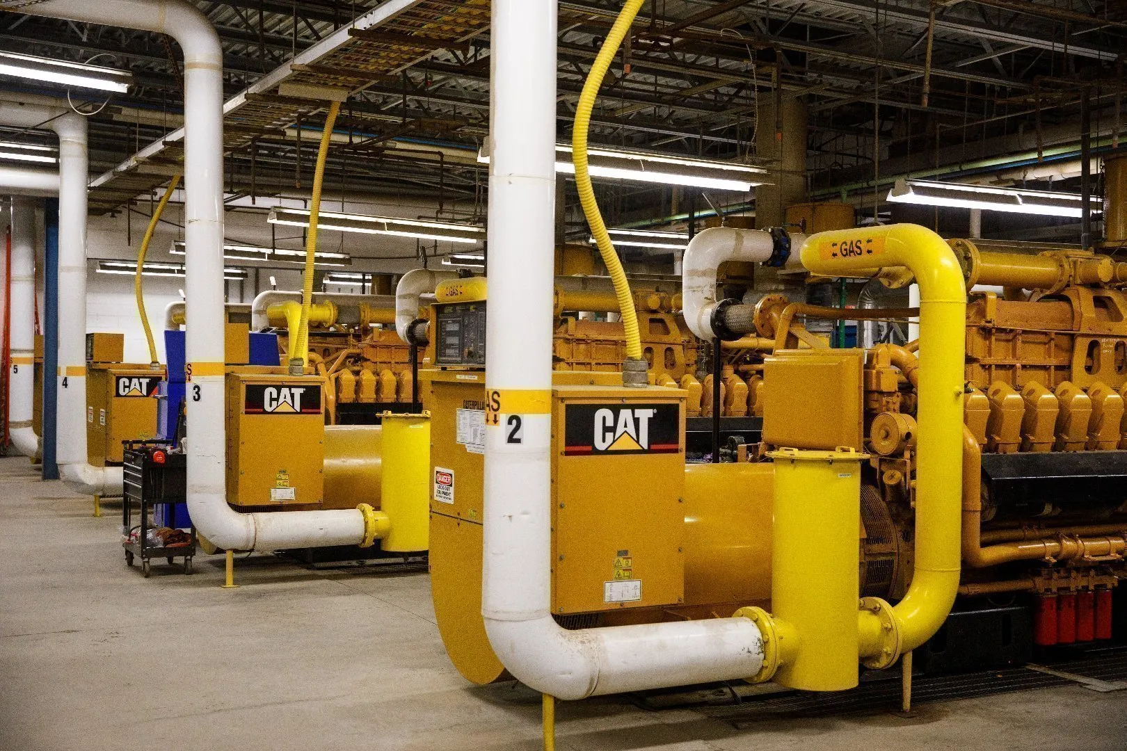 Indoor landfill gas-to-energy facility featuring multiple large yellow CAT generator engines connected to bright yellow and white piping labeled “GAS.” Overhead metal beams, ventilation ducts, and industrial lighting are visible across the ceiling, with concrete flooring throughout the space.