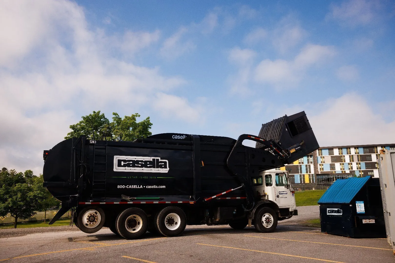 Outside a university building, a Casella front-load truck arm lifts commercial garbage can above hopper for dumping. 