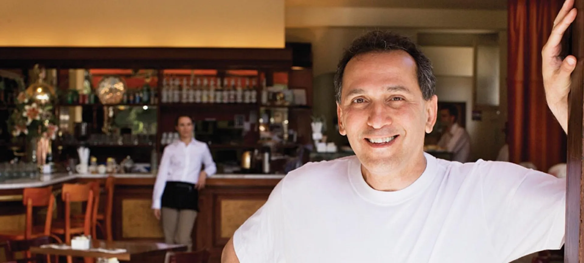 Person wearing a plain white T-shirt stands inside a restaurant or café near a wooden bar counter with shelves stocked with bottles and glassware. In the background, another individual wearing a white shirt and black apron is positioned near the bar area.