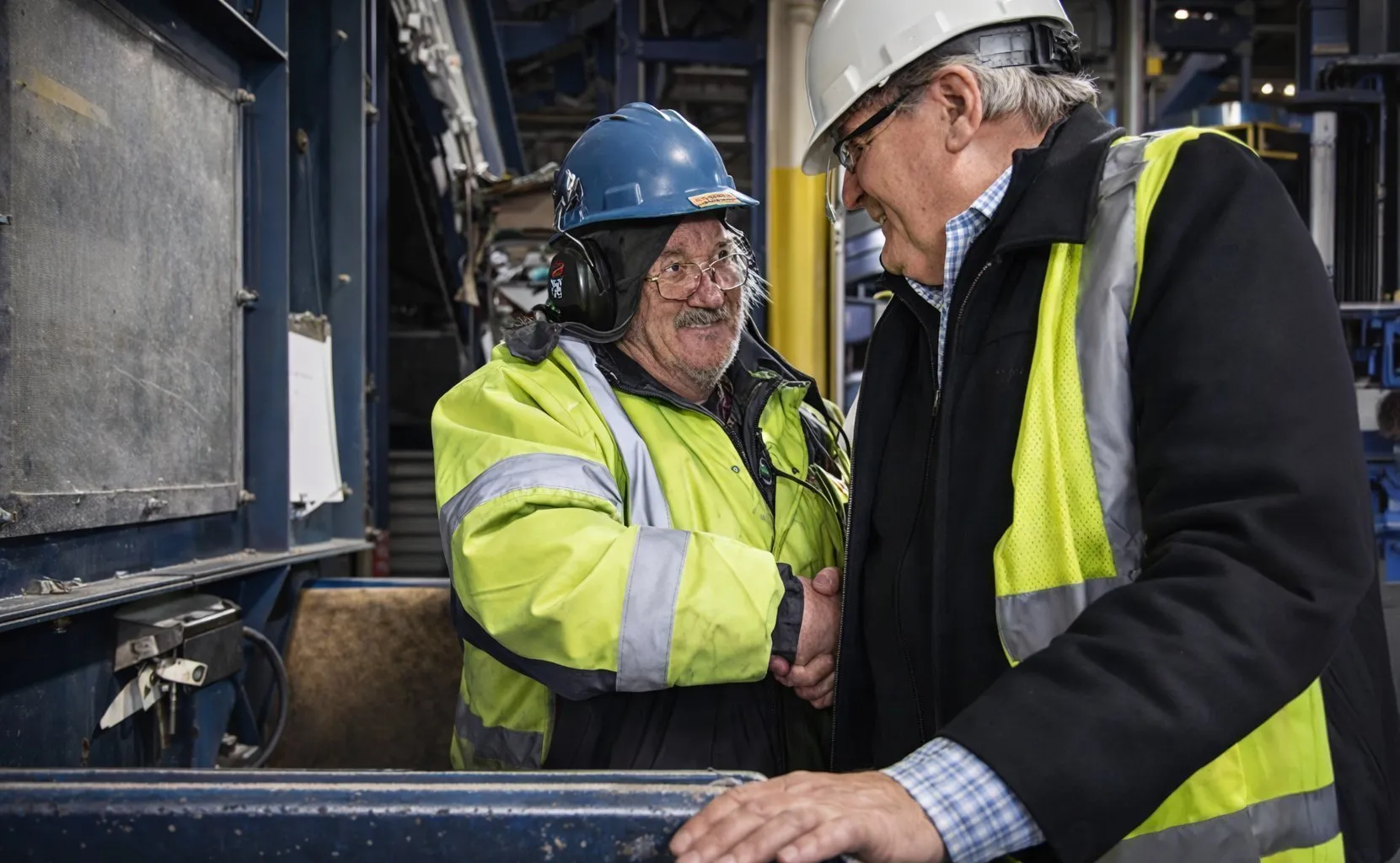 Two employees wearing hard hats and high‑visibility jackets shake hands in an industrial setting surrounded by machinery.