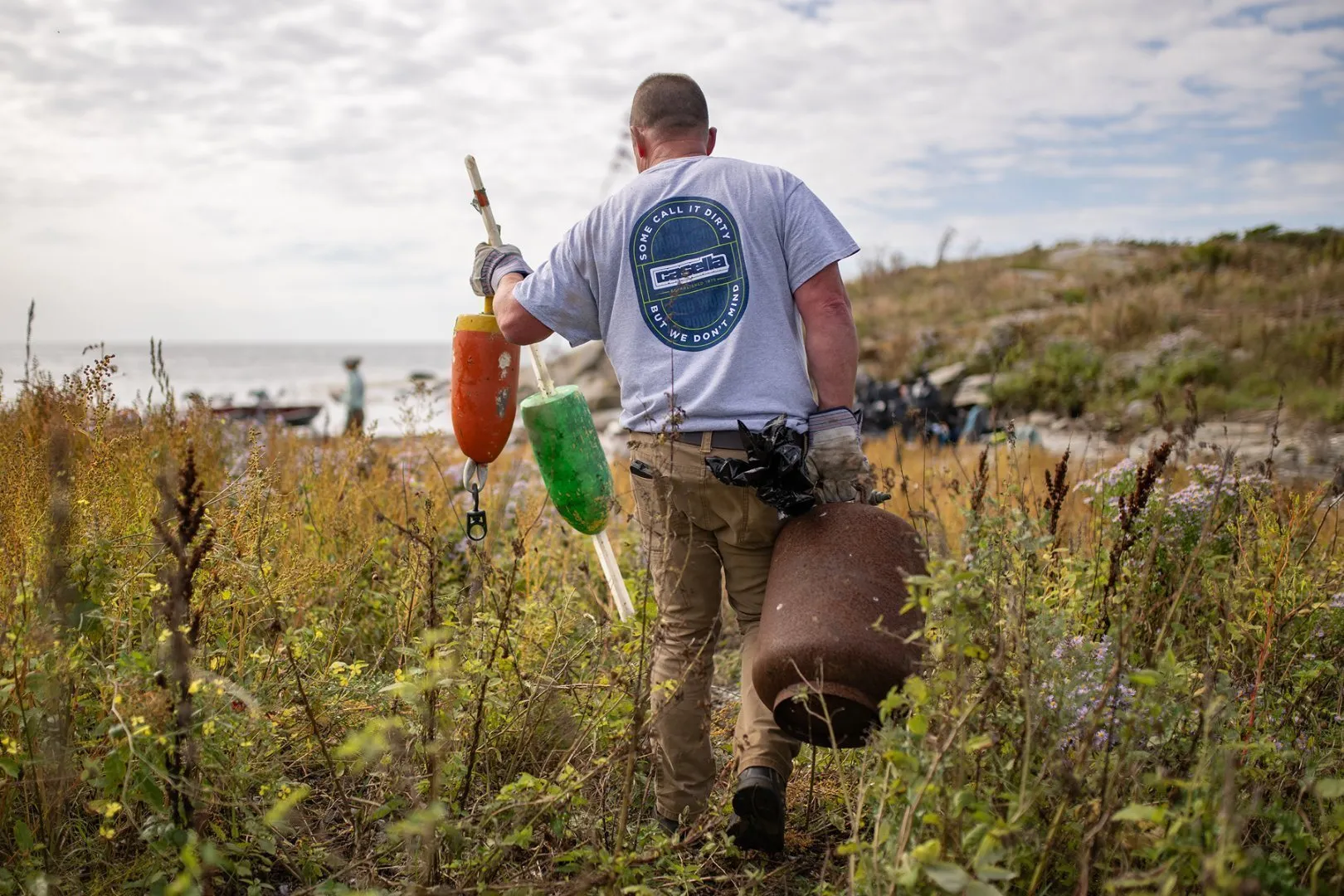 Person wearing a gray T-shirt with a Casella logo on the back walks through tall grass near a rocky shoreline, carrying three colorful buoys and marine debris. The ocean and cloudy sky are visible in the background, with boats anchored offshore.