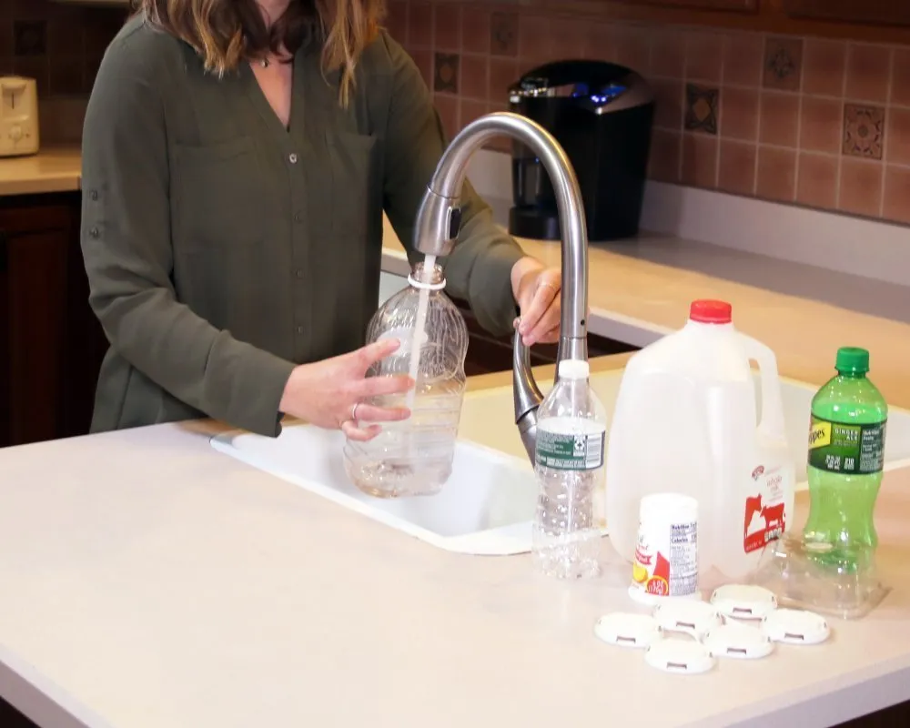 Person wearing a long-sleeve green shirt rinses a clear plastic bottle under a kitchen faucet at a white countertop sink. Other recyclable items, including a plastic water bottle, a gallon milk jug, a green soda bottle, a small white container, and several white plastic caps, are arranged on the counter. The background features a tiled backsplash, a toaster, and a coffee maker. 