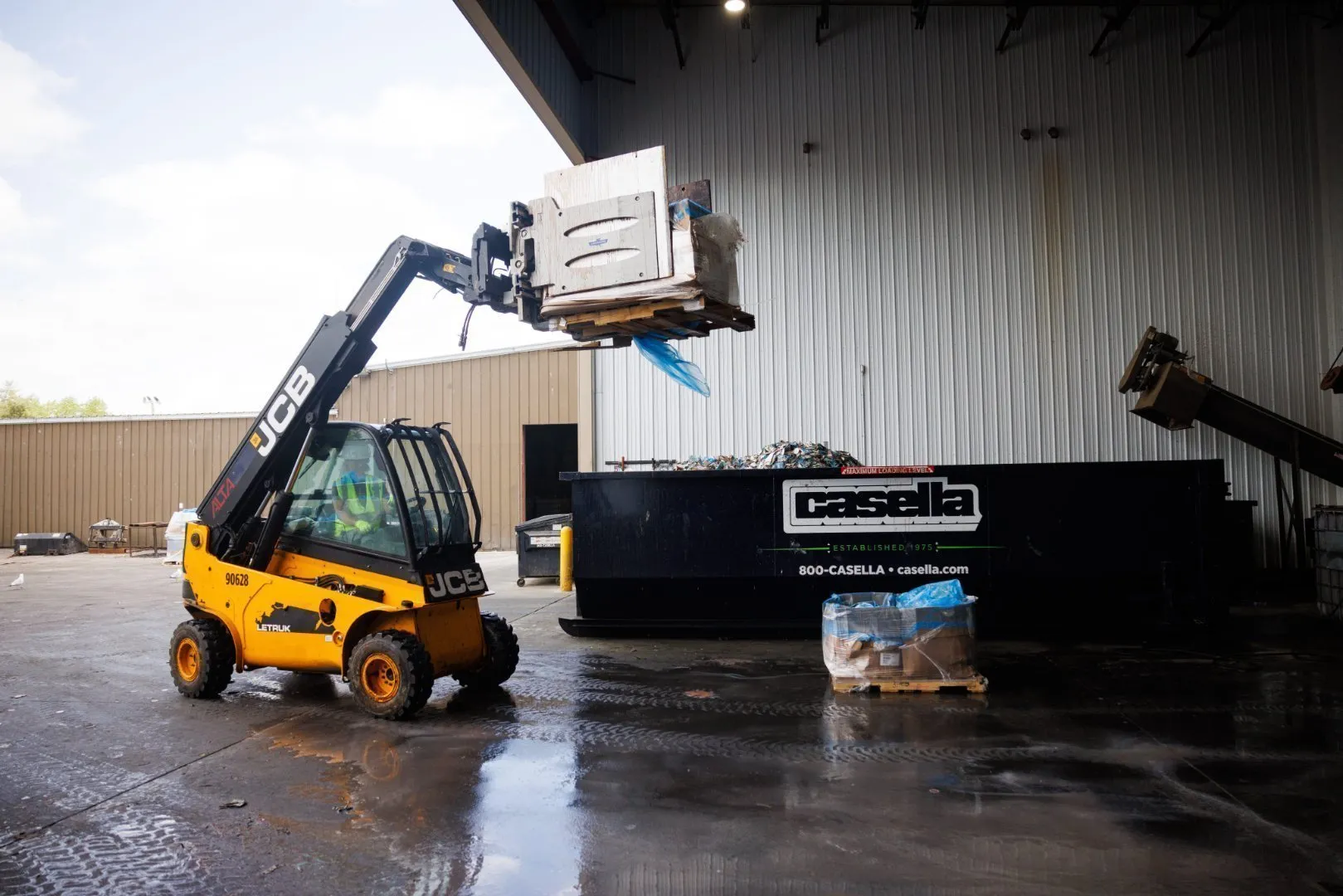 Yellow JCB telehandler lifts a large metal appliance wrapped in blue plastic inside an industrial facility, preparing to load it into a Casella roll-off dumpster. The concrete floor is wet, and additional wrapped items are stacked on a wooden pallet nearby.